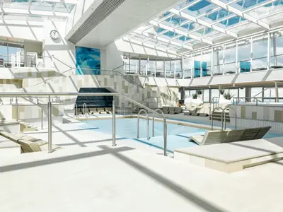 Dieses Pressefoto zeigt den lichtdurchfluteten Innenpoolbereich auf Deck 12 der Mein Schiff 3, mit einem großen Glasdach und Liegestühlen am Beckenrand. Im Hintergrund ist eine Treppe und ein weiteres Deck zu sehen.