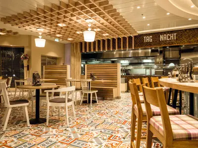 Ein heller Restaurantbereich mit beigen Wänden, Holzböden und einer Gitterdecke aus Holz an Bord der Mein Schiff 3. In der Mitte stehen runde Holztische mit weißen Stühlen. Rechts befindet sich eine Bar mit Holztheke und hohen Stühlen, darüber eine offene Küche mit dem Schriftzug "TAG & NACHT BISTRO".