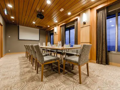 Innenansicht eines Konferenzraums an Bord der Mein Schiff 3. Ein langer Holztisch mit Stühlen, Wasserflaschen und Gläsern steht im Vordergrund. An der Wand befindet sich eine weiße Projektionsleinwand. Große Fenster auf der rechten Seite bieten einen weiten Blick auf das Meer unter einem bewölkten Himmel.