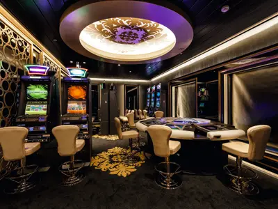 Ein luxuriöses Casino an Bord der Mein Schiff 3 mit Spielautomaten an der linken Wand und einer Gruppe von Hochstühlen um einen zentralen Spieltisch in der Mitte. Der Boden ist mit einem opulenten goldenen Muster verziert. Die Decke zeigt eine große, beleuchtete kreisförmige Leuchte mit violetten und weißen Mustern.