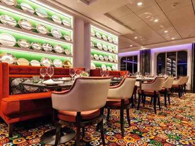 Innenansicht des Restaurants 'Surf & Turf Steakhouse' an Bord der Mein Schiff 3. Rote gepolsterte Bänke und helle Stühle an Tischen, mit beleuchteten Regalen voller dekorativer Teller im Hintergrund. Fenster mit gemusterten Vorhängen und lila Beleuchtung sind sichtbar.