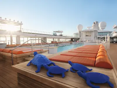 Blick auf das obere Sonnendeck der Mein Schiff 3 bei Sonnenuntergang. Ein langer, rechteckiger Pool ist von mehreren Reihen orangefarbener Liegen umgeben. Im Vordergrund sind vier blaue Schildkröten-Skulpturen zu sehen. Im Hintergrund erstrecken sich die weißen Aufbauten des Schiffes unter einem hellblauen Himmel. Die Sonne scheint hell links oben und wirft warme Strahlen über das Deck.