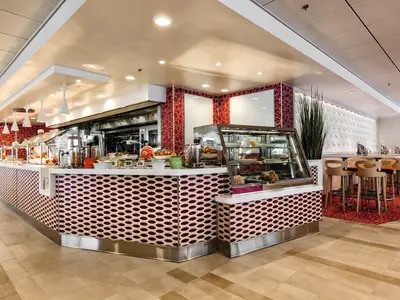 Blick in ein modernes Buffet-Restaurant auf der Mein Schiff 3. Links erstreckt sich ein langer Tresen mit Buffet-Speisen und einer Vitrine, daneben eine rote Wand mit Mustern. Rechts stehen mehrere Barhocker an hohen Tischen, der Boden ist mit einem roten Teppich ausgelegt.