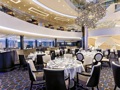 Innenansicht des zweistöckigen Restaurants Atlantic auf der Mein Schiff 3. Zahlreiche gedeckte Tische mit weißen Tischdecken und Stühle füllen den Raum. Eine glitzernde Designer-Deckenleuchte hängt von der Decke. Im Hintergrund ist eine geschwungene Treppe zu sehen.