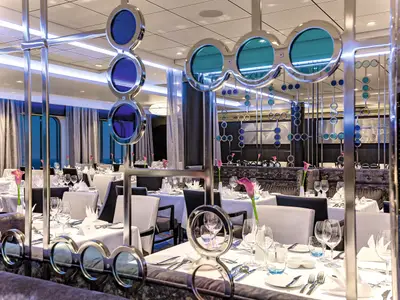 Ein helles und modern eingerichtetes Restaurant an Bord der Mein Schiff 3 mit vielen gedeckten Tischen. Helle Vorhänge, große Fenster mit Meerblick und auffällige blaue und türkise Dekoelemente prägen den Raum.