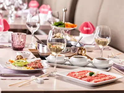 Ein festlich gedeckter Tisch auf der Mein Schiff 3 mit verschiedenen Speisen und Getränken. Im Vordergrund sind Teller mit rohem und zubereitetem Fleisch, Gemüse und zwei Gläser Weißwein zu sehen. Im Hintergrund stehen Töpfe, weitere Gläser und dekorative Elemente.