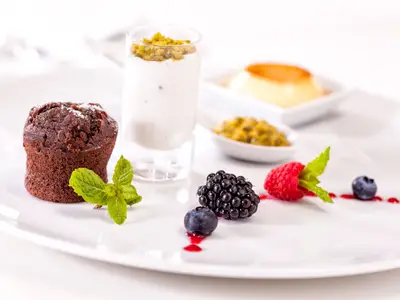 Nahaufnahme eines weißen Tellers mit verschiedenen Desserts: ein Schokoladenmuffin mit Puderzucker, ein Glas Joghurt mit Pistazien, Beeren und ein Löffel Passionsfrucht. Im Hintergrund ist eine Creme Brulee zu sehen, als Teil des Angebots auf der Mein Schiff 3.