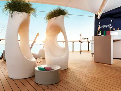 Blick von der Champagner-Bar an Bord der Mein Schiff 3 auf das offene Meer. Zwei futuristische, weiße Sitzmöbel mit Pflanzen stehen auf dem Teakholzdeck. Links ein Tisch mit zwei Weingläsern. Im Hintergrund eine Bar mit Champagnerflaschen und dem Schriftzug 'Champagner Treff'.