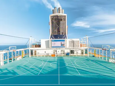 Ein Sportplatz mit Basketballkörben und Fußballtoren auf dem Deck der Mein Schiff 3. Der Platz ist türkisblau und von einem Netz sowie Glaswänden umgeben. Im Hintergrund erstreckt sich das weite, blaue Meer unter einem klaren Himmel mit einigen Wolken und einem Sichelmond.