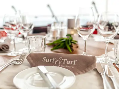 Blick auf einen elegant gedeckten Tisch im Restaurant 'Surf & Turf Steakhouse' an Bord der Mein Schiff 3. Auf der braunen Serviette liegt Besteck und ein Teller, dazu eine weitere Serviette mit 'Surf & Turf Steakhouse' Schriftzug. Im Hintergrund sind Weingläser, ein Wasserglas und eine Dekoration aus grünen Bohnen zu sehen.