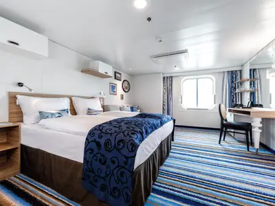 Ein helles Schlafzimmer an Bord der Mein Schiff 3 mit einem großen Doppelbett, einer blauen Tagesdecke mit Muster und einem passenden gestreiften Teppich in Blau- und Brauntönen. Rechts ist ein Schreibtisch mit Stuhl und ein Fenster zu sehen.