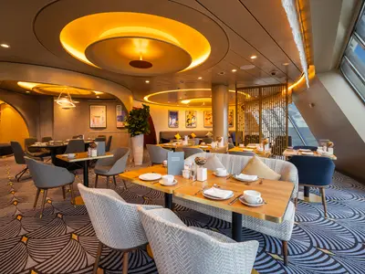 Innenansicht des Restaurants an Bord der Mein Schiff 3 mit zahlreichen Tischen und Stühlen, warmem Licht und großen Fenstern mit Blick auf das Meer. Der Teppich hat ein Wellenmuster.