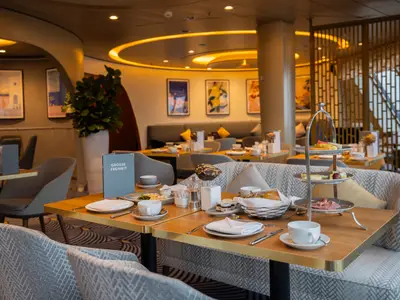 Ein liebevoll gedeckter Tisch mit Frühstück und einer Etagere mit süßen und herzhaften Speisen steht an Bord der Mein Schiff 3 im Restaurant „Grosse Freiheit“. Im Hintergrund sind weitere Tische und Kunst an den Wänden zu sehen.