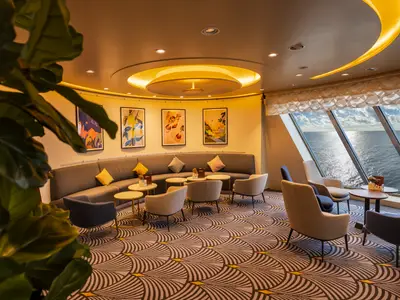 Ein breiter Blick in die stilvolle, helle Lounge der Mein Schiff 3. Im Vordergrund sind Pflanzen zu sehen. Eine geschwungene graue Bank mit bunten Kissen und mehreren Tischen dominiert den linken Bereich. Der rechte Teil bietet Sessel und weitere Tische mit weitem Blick auf das sonnige Meer durch große Fenster.
