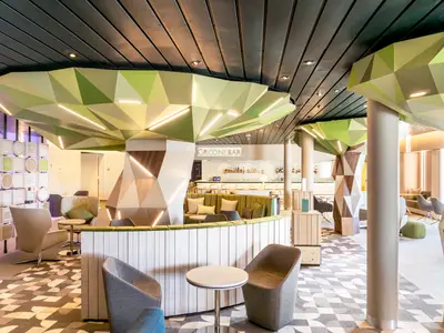 Blick in die moderne "Groöne Bar" Lounge der Mein Schiff 3. Die Decke ist mit geometrischen, grün getönten Formen verziert, die in der Mitte der Bar leuchtende Lichtstreifen aufweisen. Im Vordergrund sind bequeme Sessel und Tische zu sehen, während im Hintergrund eine lange, beleuchtete Bar mit Hockern und Getränkeflaschen erkennbar ist. Der Boden ist mit einem geometrischen Musterteppich ausgelegt. Seitlich stehen weitere Sitzgruppen und Säulen mit geometrischer Verkleidung.