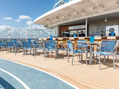 Blick auf das obere Deck der Mein Schiff 3 mit einer Outdoor-Bar und vielen leeren Tischen und Stühlen mit blauen Sitzflächen. Im Hintergrund ist eine Glasbrüstung und der blaue Himmel zu sehen. Ein Barkeeper steht bereit.