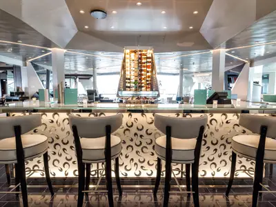 Blick auf die moderne Bar an Bord der Mein Schiff 3. Im Vordergrund stehen vier Barhocker mit cremefarbenen Polstern. Die Bar selbst ist mit einem beleuchteten Muster verziert. Dahinter, als zentrales Element, befindet sich ein hohes, futuristisch anmutendes Weinflaschenregal, das eine Vielzahl von Flaschen präsentiert. Große Fenster im Hintergrund bieten einen Ausblick auf die Umgebung.