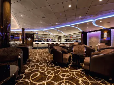 Innenansicht der TUI Bar an Bord der Mein Schiff 3 mit braunen Sesseln und runden Tischen auf einem gemusterten Teppich. Im Hintergrund befindet sich eine gut bestückte Bar mit Spiegeln und Flaschen, beleuchtet von blauen und violetten Akzentlichtern.