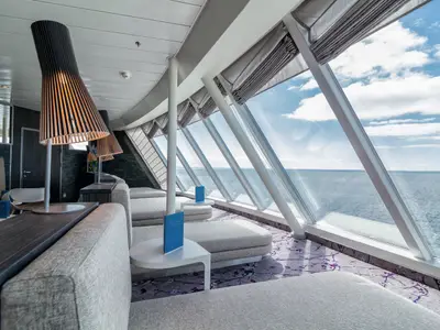 Innenansicht eines stilvollen Loungebereichs auf der Mein Schiff 3 mit geneigten Panoramafenstern, die einen weiten Blick auf das offene Meer und den blauen Himmel ermöglichen. Mehrere bequeme hellgraue Liegen mit kleinen Beistelltischen und blauen Menükarten laden zum Verweilen ein. Die Decke ist hell und mit modernen Holzlamellen-Pendelleuchten versehen, die ein warmes Licht abgeben. Der Boden ist mit einem gemusterten Teppich in Grau- und Violetttönen ausgelegt. Im Hintergrund sind dunkle Holzwände und weitere Sitzgelegenheiten zu sehen. Die Atmosphäre ist ruhig und einladend.