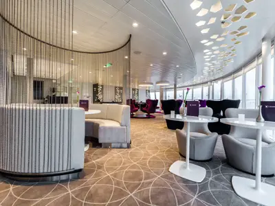 Ein moderner Lounge-Bereich an Bord der Mein Schiff 3 mit geschwungener Decke, großen Fenstern und verschiedenen Sitzbereichen. Links eine runde Sitzbank mit Perlenvorhang, rechts Tische mit grauen Sesseln, im Hintergrund violette Sessel. Die Decke ist mit dekorativen Lichtelementen versehen.