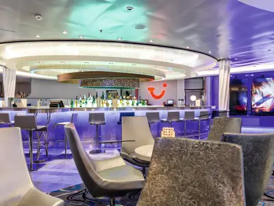 Innenansicht einer modernen Bar an Bord der Mein Schiff 3. Eine lange, beleuchtete Bar mit vielen Flaschen und Barhockern. Im Hintergrund sind große Bildschirme und Sitzgelegenheiten zu sehen. Die Beleuchtung ist überwiegend lila.