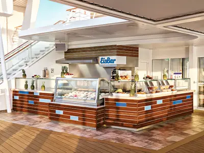 Detailaufnahme des EisBar-Buffets auf dem Sonnendeck der Mein Schiff 3. Ein moderner Stand mit Holzakzenten und blauen Paneelen bietet eine Auswahl an Eis und frischen Früchten. Mehrere Ananas dienen als Dekoration. Im Hintergrund eine Treppe, die zu weiteren Decks führt.
