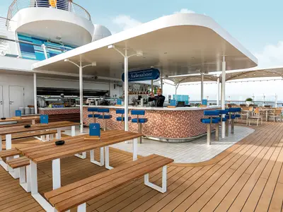 Ein Barkeeper mixt Drinks an der Aussenalster Bar der Mein Schiff 3. Zahlreiche Tische und Bänke laden zum Entspannen an Deck ein. Im Hintergrund ist das Meer sichtbar.