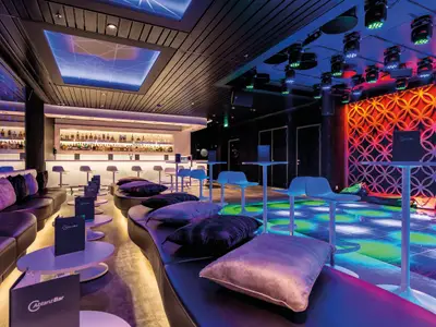 Ein luxuriöser Barbereich auf der Mein Schiff 3 mit einer langen Theke, gemütlichen Sofas, stilvollen Tischen und einer farbig leuchtenden Tanzfläche. Die Decke ist dunkel mit Beleuchtungssystemen, und eine dekorative Wand mit runden Mustern in Rot- und Orangetönen akzentuiert den Barbereich.