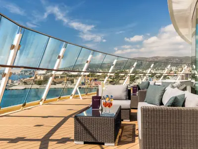 Sonniger Tag an Bord der Mein Schiff 3, mit Blick von einer Terrasse mit Rattanmoebeln, Holzdeck und Glasgelaender auf eine Kuestenstadt mit Gebirge im Hintergrund.