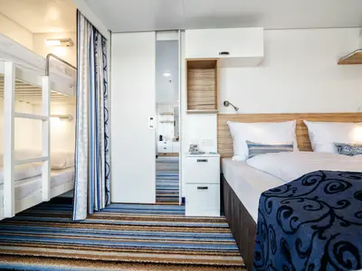 Innenansicht einer geräumigen Kabine der Mein Schiff 3 mit einem Doppelbett, einem Nachttisch mit Schubladen, und einem Etagenbett hinter einem gestreiften Vorhang. Ein Spiegel reflektiert einen weiteren Raum.