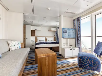 Innenansicht einer hellen Suite an Bord der Mein Schiff 3 mit einem grauen Sofa, einem Holztisch, einem Doppelbett im Hintergrund und einem Fernseher. Große Fenster bieten einen weiten Blick auf das Meer. Ein blauer Sessel mit floralem Muster steht rechts.