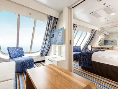 Innenansicht einer Kabine auf der Mein Schiff 3 mit bodentiefen Fenstern, die Meerblick bieten. Die Kabine ist in Weiß, Blau und Holzfarben gehalten. Im Vordergrund ein Wohnbereich mit Sofa, Couchtisch und einem blauen Sessel mit Blattmuster. Dahinter ein weißer Schrank mit Fernseher und ein angrenzender Schlafbereich mit Bett, das eine dunkelblaue Decke mit Muster hat.