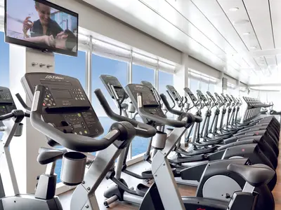 Blick in den hellen Fitnessraum der Mein Schiff 3 mit raumhohen Fenstern, die einen weiten Meerblick bieten. Mehrere Reihen von Life Fitness Cardiogeräten, darunter Crosstrainer und Fahrradergometer, stehen bereit. An der vorderen Säule hängt ein Fernseher, der eine Unterhaltungsszene zeigt.