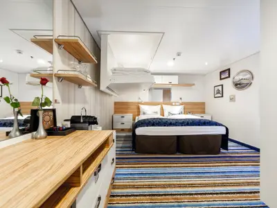 Ein helles Schlafzimmer auf der Mein Schiff 3. Ein Doppelbett mit blauer Tagesdecke dominiert den Raum. Darüber ein ausklappbares Bett. Rechts an der Wand hängen Seefahrtsbilder. Links ein Waschtisch mit Kaffeemaschine und roten Rosen, daneben Regale.