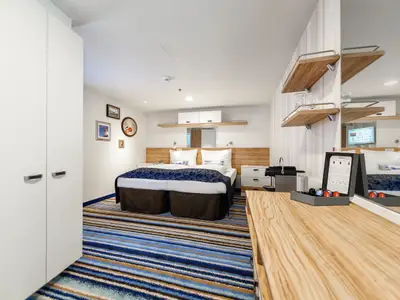 Blick in eine moderne Kabine der Mein Schiff 3 mit einem Doppelbett in der Mitte, umgeben von Holz- und Weißtönen. Links ein weißer Schrank, rechts eine Kaffeemaschine und ein Spiegel. Der Boden ist mit einem blau-braun gestreiften Teppich ausgelegt.