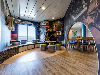 Ein stylischer Loungebereich an Bord der Mein Schiff 3 mit maritimen Blick, Sitzgelegenheiten am Fenster, einem runden Tisch mit Hockern und einer Wandgestaltung mit Autoteilen.