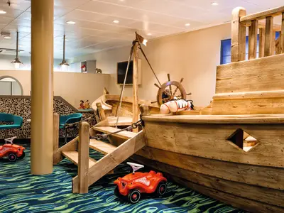 Ein Detailansicht des Kinder- und Jugendbereichs "Insel der Seeräuber" an Bord der Mein Schiff 3. Man sieht ein hölzernes Spielschiff mit Steuerrad, eine Leiter und zwei rote Bobbycars auf einem Teppich mit blau-grünem Wellenmuster.