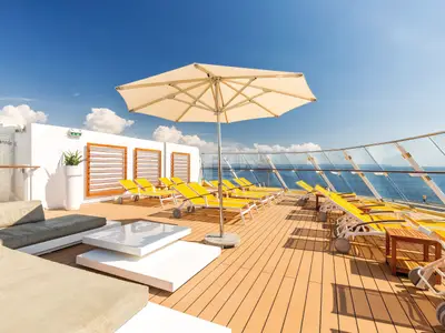 Sonnendeck der Mein Schiff 3 mit vielen gelben Liegestühlen unter einem großen beigen Sonnenschirm. Im Hintergrund ist das blaue Meer und ein heller Himmel mit einigen Wolken zu sehen.
