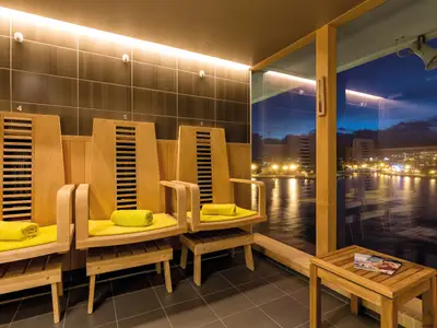 Innenansicht eines Raumes an Bord der Mein Schiff 3, der drei helle Holzliegen mit gelben Handtüchern zeigt. Durch eine große Glaswand blickt man auf eine erleuchtete Stadt und das Meer bei Dunkelheit.