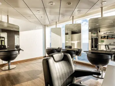 Ein heller Friseursalon an Bord der Mein Schiff 3 mit Holzboden und drei modernen Friseurstühlen. Große Fenster bieten einen weiten Blick auf das offene Meer. An der Wand hängen Spiegelschranke, und in der Reflexion ist ein 'Color Bar' zu sehen. Die Decke ist mit quadratischen Paneelen und Einbauleuchten versehen.