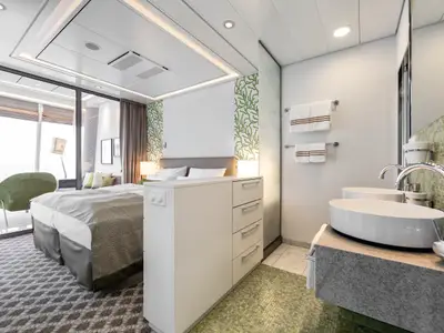 Innenansicht einer geräumigen Suite an Bord der Mein Schiff 3. Man sieht ein Bett, eine Sitzecke am Fenster mit Meerblick, einen Waschtisch mit zwei Aufsatzwaschbecken und einen Spiegelschrank. Die Wände sind hell, mit grünen Korallenmuster-Elementen.