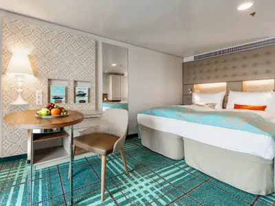 Detailaufnahme einer hellen Kabine an Bord der Mein Schiff 3. Man sieht das Doppelbett mit türkiser Tagesdecke, dahinter eine gepolsterte Kopflehne und eine gemusterte Tapete. Links steht ein kleiner runder Tisch mit Obstteller und ein Stuhl, darüber eine Lampe und Regale mit Broschüren.