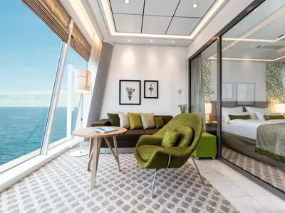 Ein moderner Wohn-/Schlafbereich an Bord der Mein Schiff 3. Große Fensterfronten bieten Meerblick. Ein braunes Sofa und ein grüner Sessel laden zum Entspannen ein. Ein Doppelbett ist durch eine Glaswand abgetrennt.