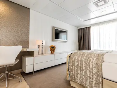Eine helle und modern eingerichtete Kabine an Bord der Mein Schiff 3. Man sieht einen Schreibtisch mit Stuhl, einen Flachbildfernseher an der Wand, eine lange Anrichte und ein Bett mit gemusterter Decke. Große Fensterfronten bieten viel Tageslicht und den Blick aufs Meer.