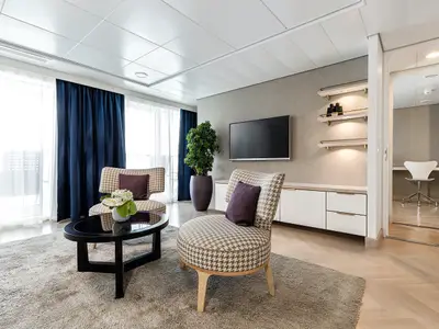 Ein stilvoller Wohnbereich an Bord der Mein Schiff 3 mit zwei gemusterten Sesseln, einem Couchtisch mit Blumen und einem Fernseher an der Wand. Rechts ist ein offener Durchgang zu einem Arbeitsbereich mit Schreibtisch und Stuhl zu sehen. Der Raum hat helle Holzböden und eine Tür zu einem Balkon mit Vorhängen.