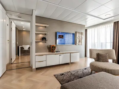 Ein helles Zimmer an Bord der Mein Schiff 3 mit Holzboden und strukturierten Wänden. Es gibt einen Fernseher, ein Sofa, einen Sessel und einen Blick in einen angrenzenden Arbeitsbereich mit Schreibtisch und Stuhl.