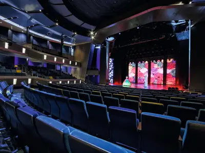 Blick von den Rängen auf das leere Theater der Mein Schiff 3. Blaue und gelbe Sitze führen zur Bühne, auf der eine bunte Projektion und zwei Figuren in Kostümen zu sehen sind.
