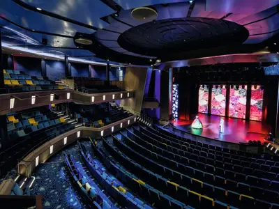 Blick von oben auf das moderne Theater an Bord der Mein Schiff 3. Reihen von blauen und gelben Sitzen füllen den Raum. Auf der Bühne stehen zwei Schauspieler vor einer großen Videoleinwand mit farbigen Motiven.