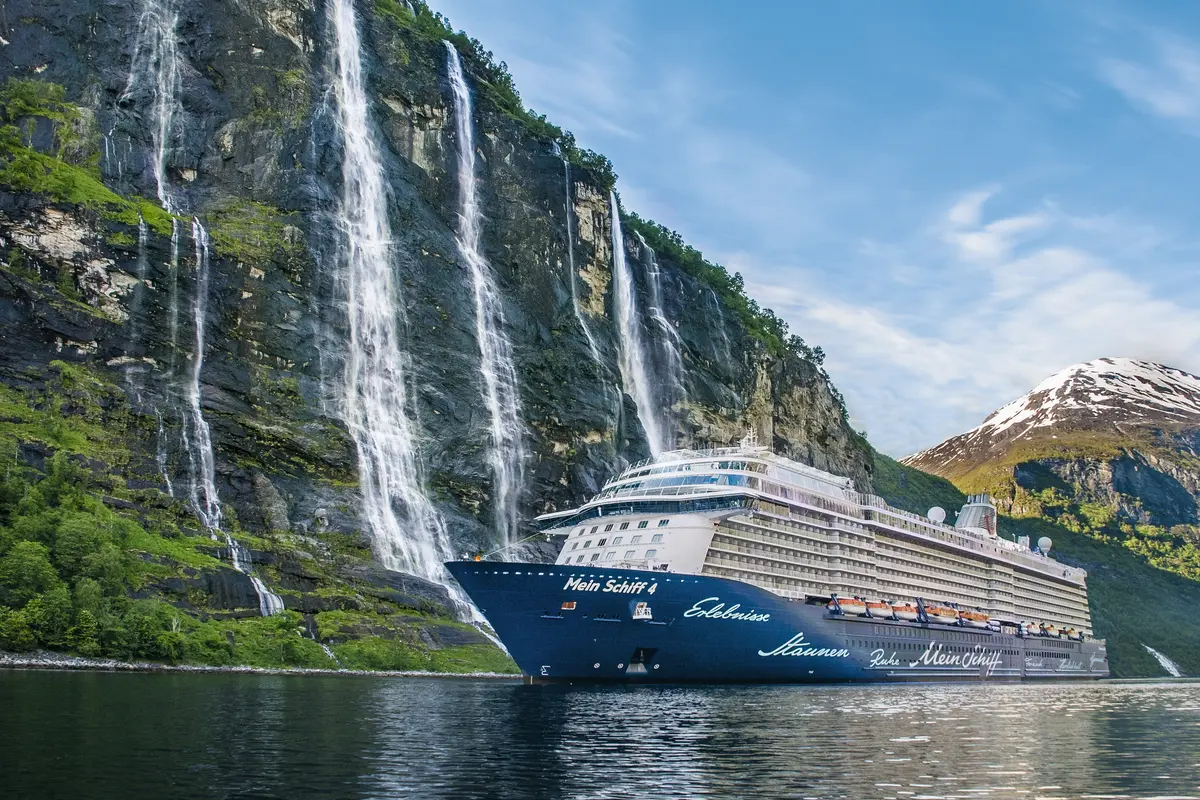 Die Mein Schiff 4 fährt durch einen tiefen Fjord. Links sind steile Felswände mit mehreren Wasserfällen zu sehen, rechts erheben sich grüne Berge mit schneebedeckten Gipfeln unter blauem Himmel.