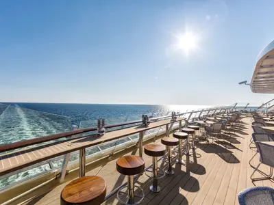 Ein sonniges Deck am Heck der Mein Schiff 4 mit einer langen Bar mit Hockern und zahlreichen Tischen und Stühlen. Das Schiff hinterlässt eine Schaumspur im blauen Meer.